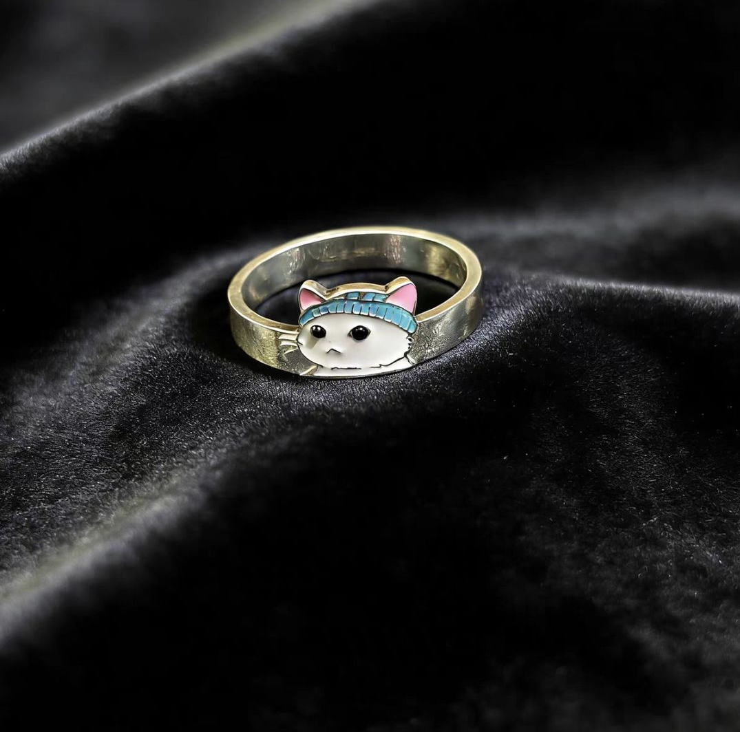 A clean product shot of the 925 sterling silver ring featuring a white enamel cat with a blue beanie, placed on black velvet.