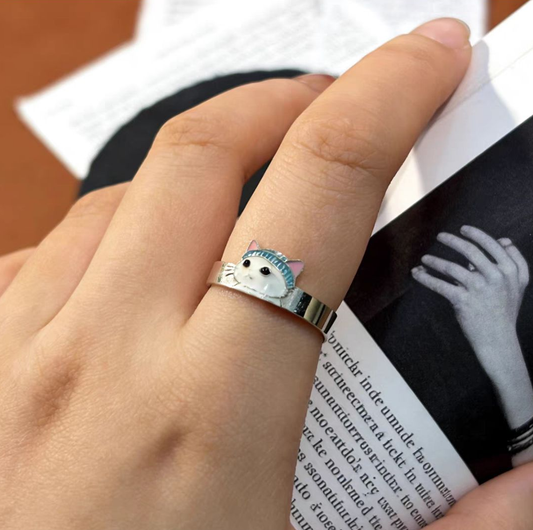 A 925 sterling silver ring with a white enamel cat in a blue beanie hat, worn on a model's finger next to a coffee cup.